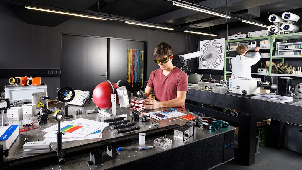 Laboratorio con apparecchiature tecniche e strumenti di misura su tavoli, due persone in abbigliamento da lavoro, una utilizza dispositivi, l'altra è seduta al tavolo.