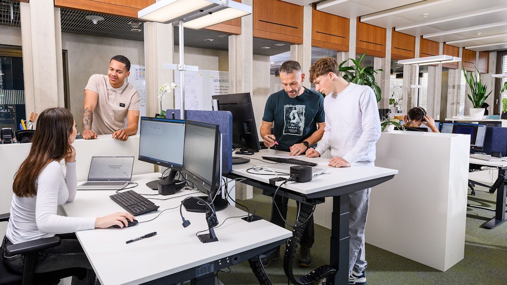 Più persone in un ufficio moderno con computer su scrivanie bianche, piante e pannelli di legno al soffitto.