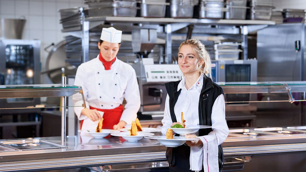 Cameriera con camicia bianca e gilet nero che porta un piatto con gamberi e contorno in cucina professionale.