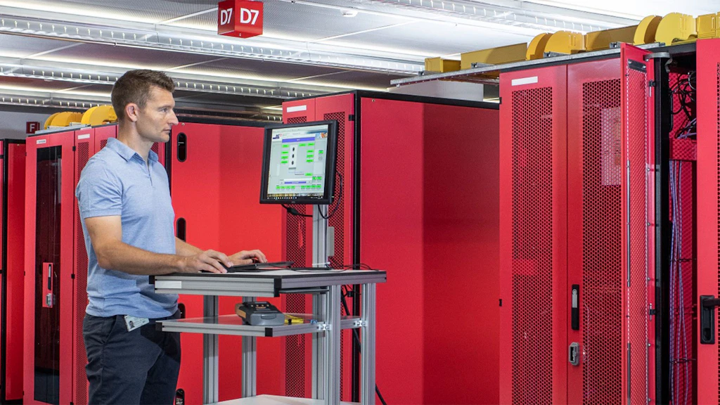 Uomo in piedi a una postazione mobile con tastiera e monitor davanti a rack di server rossi in un centro dati.