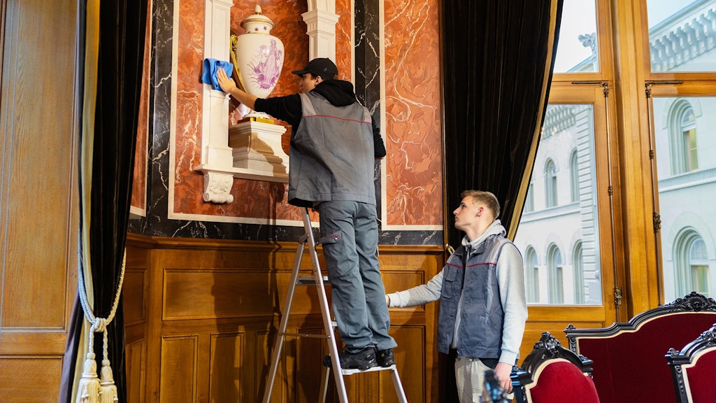 Persona su una scala che pulisce un vaso bianco in una stanza ornata con boiserie e grandi finestre.